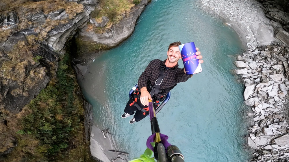 An adventurous man in his twenties hangs from a canyon swing holding a chuffed gifts container in his left hand. He is dangling over a body of water